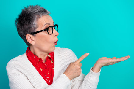 Mature woman with glasses wearing red christmas shirt and white cardigan points to her open hand against teal background in a festive lifestyle portraitの写真素材