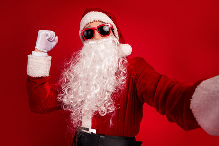 Santa in red suit with white beard posing in sunglass hat against bright red background festive Christmas themed photo shootの写真素材