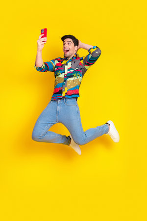 Joyful young male model jumps with phone against a bright yellow background wearing a colorful print...