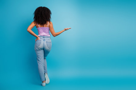 Young girl in lilac top and jeans stands against blue background posing with hand out and back turned to cameraの写真素材