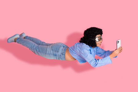 Young woman wearing goggles holding a smartphone while pretending to fly in front of a pink...