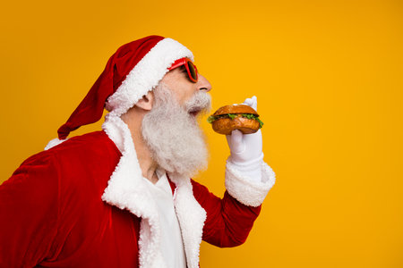 Festive Santa Claus enjoying a burger while celebrating Christmas against vibrant yellow backgroundの写真素材