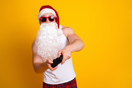 Playful Santa with white beard and red hat holds a game controller in a bright yellow studio...