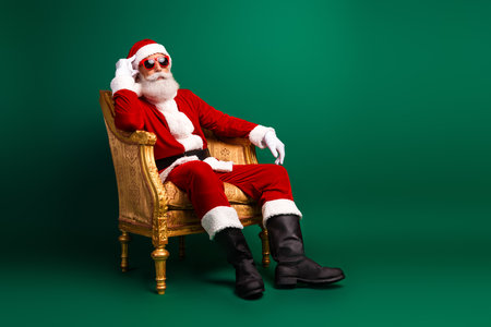 Stylish Santa Claus in a trendy suit relaxing on an ornate chair with a green festive background spreading cheerful vibesの写真素材