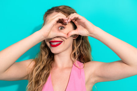 Young woman forms a heart frame with her hands smiling brightly against a turquoise backdrop for fashion lifestyle and fun stock photoの写真素材