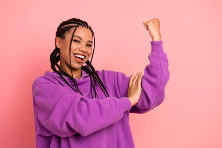 Portrait of a cheerful young woman flexing arm muscle, wearing purple hoodie, displaying confidence and positivityの写真素材