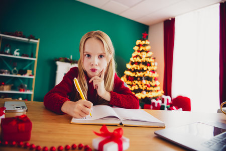 Playful girl at her desk writes in a cozy home during christmas time with a bright tree and festive mood as soft lights glow around gifts and decorations creating warmth and joyの写真素材