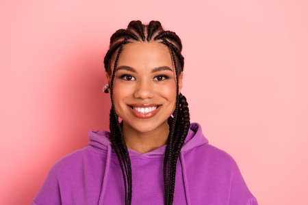 Smiling young woman with braids wearing a purple hoodie posing in front of a pink background
