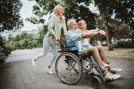 Family joy in the park as a granddaughter helps push grandpa in a wheelchair with a smiling daughter nearbyの写真素材
