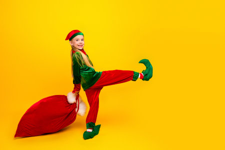 Brightly dressed child wearing holiday elf costume posing with red sack against vibrant yellow backgroundの写真素材