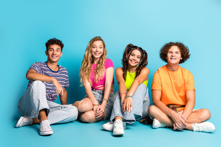 Bright teens friends sitting together in a studio with a blue backdrop smiling and enjoying colorful casual outfitsの写真素材