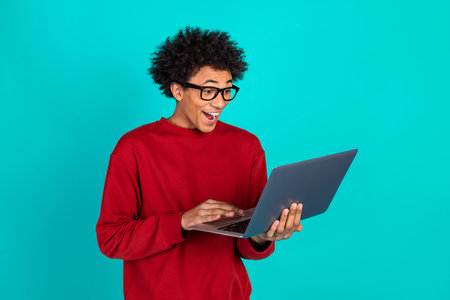 Young man in red sweater using laptop with surprised expression against bright teal background for...