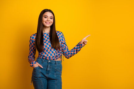 Young stylish Latina student woman stands against a bright yellow background smiling and pointing to the right showcasing casual chic fashion and confident modern lifestyleの写真素材