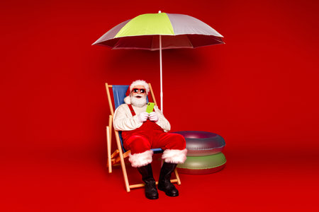 Santa relaxing under a colorful umbrella holding a phone during a festive Christmas scene with playful props and seasonal vibesの写真素材