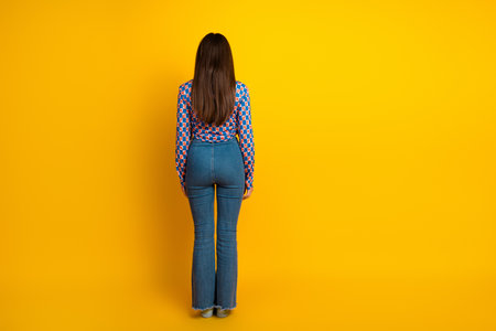Young woman with long brunette hair wearing a patterned top and jeans stands with her back to the camera against yellow backgroundの写真素材