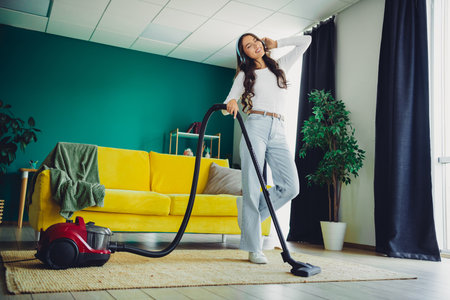 Young woman enjoys cleaning at home in a bright living room with a yellow couch using a vacuum cleaner for a relaxed modern home lifestyleの写真素材