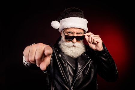 Cool Santa in leather jacket points at you wearing sunglass and hat for festive holiday portraitの写真素材