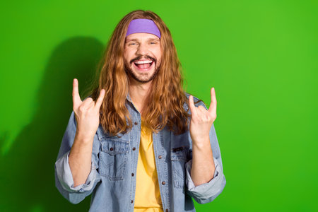 Portrait of a cheerful long-haired man wearing denim, making rock hand gestures against a vivid...