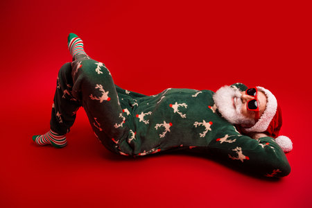 Relaxing Santa in festive green reindeer onesie wearing red sunglass on a bold red background dreaming of holiday joyの写真素材