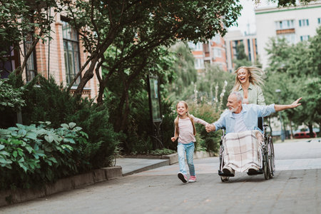 Joyful family outing as granddaughter and mother push grandpa in wheelchair through a green park beside a bustling city streetの写真素材