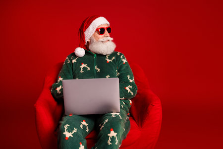 Santa relaxes in festive pajama with laptop in a cozy Christmas moment against a red background for...