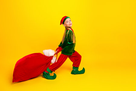 Smiling young girl in elf costume holding Santas sack against vibrant yellow background during...
