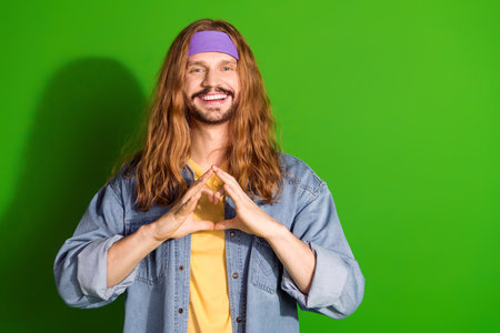 Young man with long hair smiling and forming a hand gesture in front of a vibrant green backgroundの写真素材