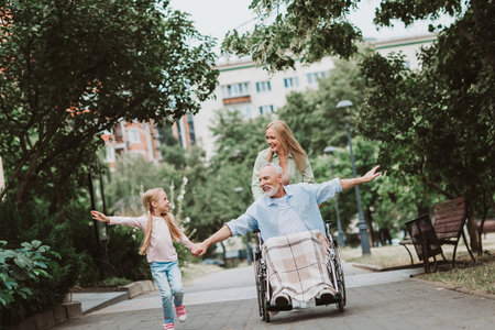 Joyful family outing in the park as grandpa in wheelchair shares a smile with granddaughter and daughter guiding them along a sunny nature pathの写真素材