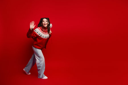 Young woman in a festive red Christmas sweater waves cheerfully against a vivid red background perfect for holiday campaigns and seasonal fashion visualsの写真素材