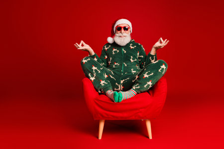 Festive Santa in green reindeer pajamas sits cross legged on a red chair against a red background wearing sunglasses for holiday cheerの写真素材
