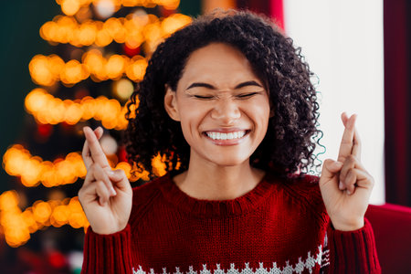 Cheerful woman in a red sweater crosses fingers by a warmly lit Christmas tree and festive decorations in a cozy home holiday settingの写真素材