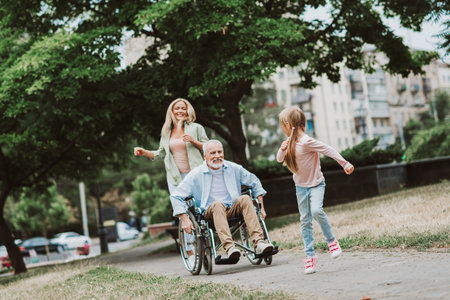 Playful moment in the park with grandpa in a wheelchair smiling as daughter runs beside him and family shares love and careの写真素材