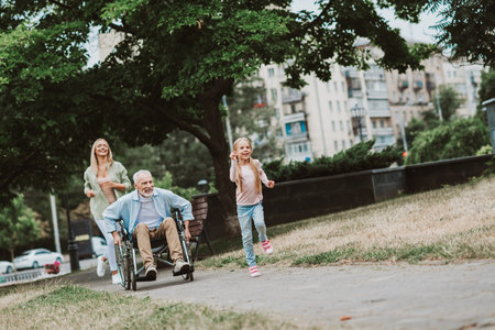 joyful family day in the park with grandpa in wheelchair running and smiling while daughter and...