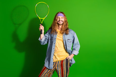 Young man with long hair in colorful retro outfit holding tennis racket and smiling against a...
