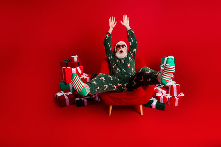 Santa in pajama raises hands amid stacked gifts in bold red studio for festive season family celebration moodの写真素材