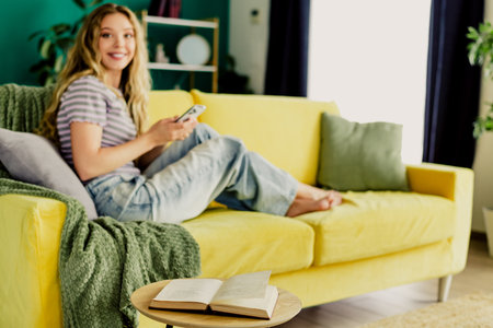 Relaxed woman at home lounging on a bright yellow sofa with a book and phone in a cozy living room settingの写真素材