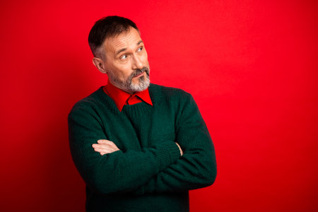 A playful guide for a mature man in a green sweater posing with crossed arms against a bold red background inviting holiday humor and festive vibes for christmas campaignsの写真素材