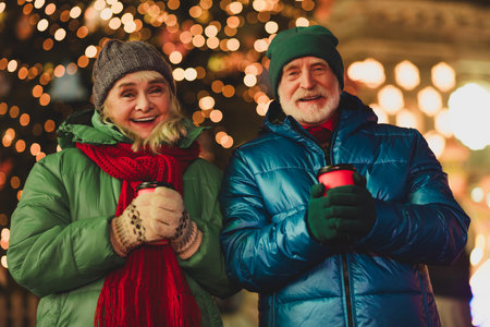 Funny warm moment outdoors with a couple of grandparents smiling holding cups in festive city lights and winter coatsの写真素材