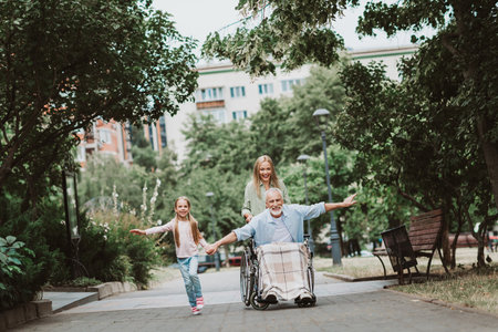 Joyful family pushes grandpa in wheelchair through a sunny park with daughter and mother walking beside smiling in a moment of care and togethernessの写真素材