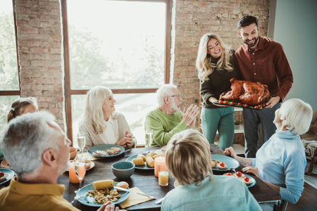 Family gathers for a festive Thanksgiving feast as a couple presents a roasted turkey to delighted seniors and grandchildren in a warm brick dining roomの写真素材