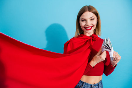 Stylish young woman with a vibrant red top holding banknotes, posing against a bright blue background with a joyful smile.の写真素材