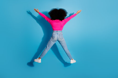 Cheerful young woman with curly hair stretching arms upward showcasing vibrant style on a blue backgroundの写真素材