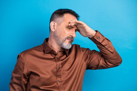 Executive man covers his eyes in a thoughtful pose wearing a brown shirt against a blue backdrop for a modern business photoshootの写真素材