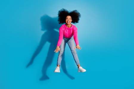Young woman in pink sweater and jeans jumping joyfully against a blue background with stylish and...