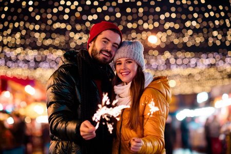Playful couple in warm winter coats light up the night with sparklers under festive lights at a cozy...
