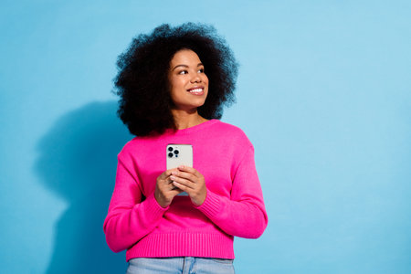 Cheerful woman in pink sweater holding a smartphone, smiling and looking away with a playful expression, blue backgroundの写真素材