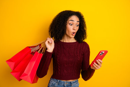 Young diverse woman with red sweater carries shopping bags and checks her phone against a bright yellow background in a casual stylish fashion momentの写真素材