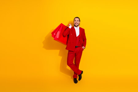 Playful stylish man in bold red suit carries shopping bags against a bright yellow backdrop for a vibrant fashion advertisementの写真素材