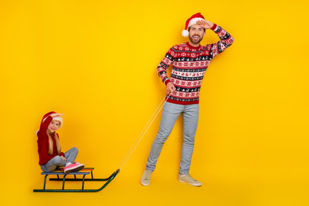 Father pulling daughter on sled wearing festive outfits on yellow backdrop, celebrating Christmas...