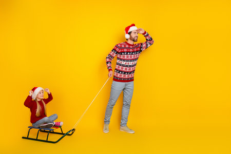 Father and Daughter Celebrating Christmas in Festive Attire with a Sled Having Fun Together on a Yellow Backgroundの写真素材